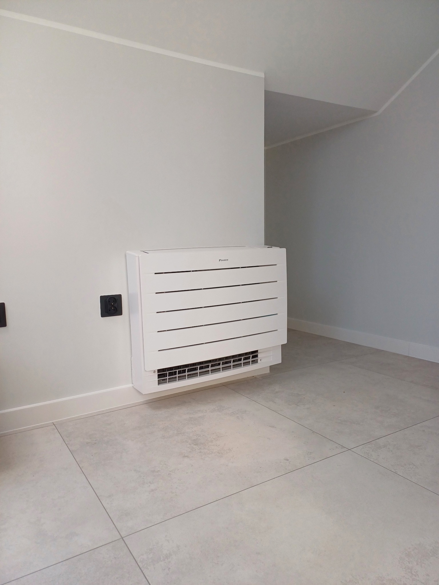 Biała, przypodłogowa jednostka klimatyzacji Daikin na tle szarej ściany i jasnej podłogi. Widoczne gniazdka elektryczne. Minimalistyczny design.