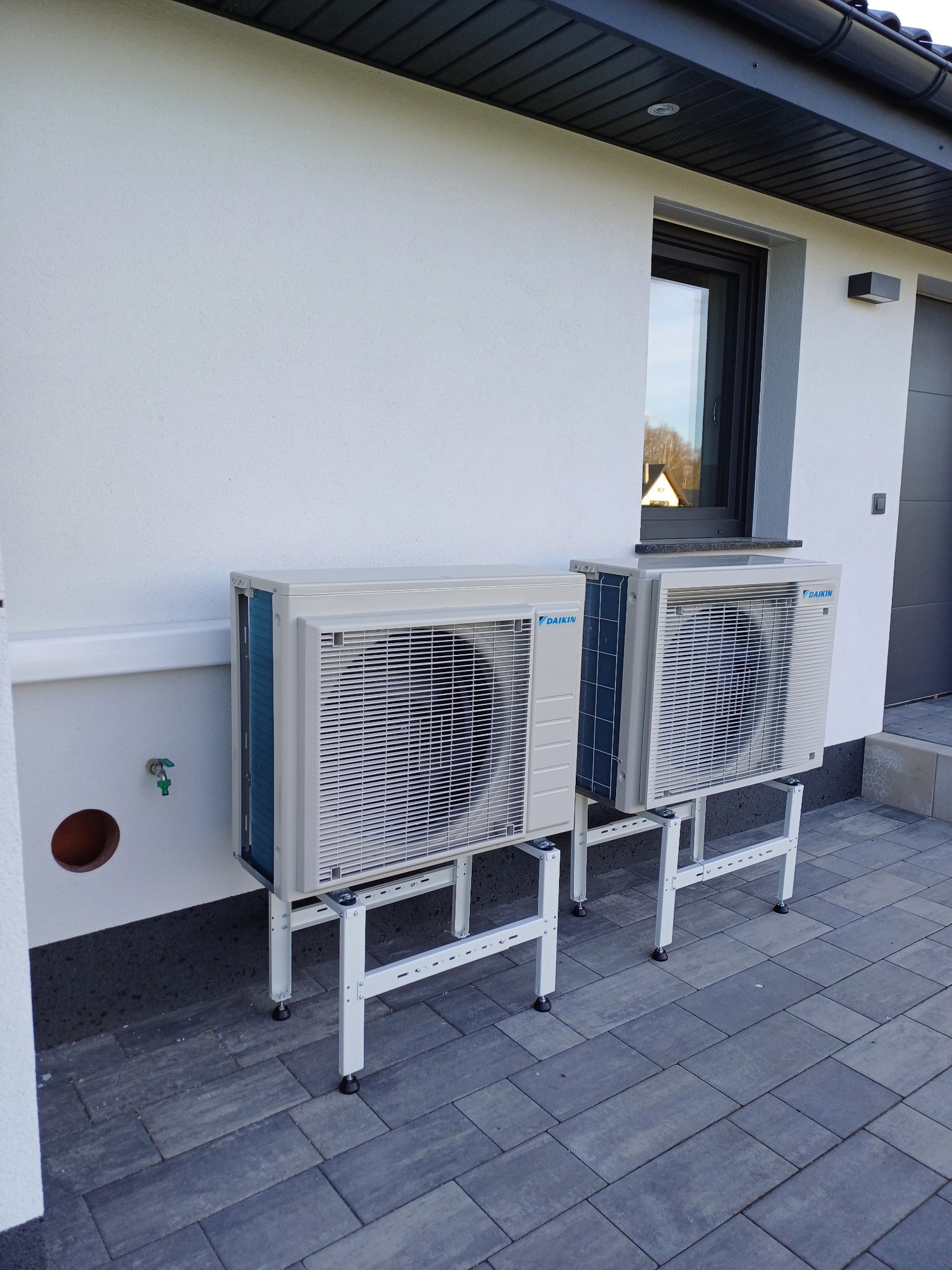 Dwa klimatyzatory Daikin zamontowane na zewnątrz budynku na białych, metalowych stelażach z regulowanymi nóżkami, na tle szarej kostki brukowej i białej ściany.