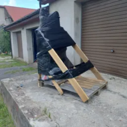 Element zabezpieczony czarną folią i drewnianą konstrukcją, prawdopodobnie do transportu lub przechowywania, na zewnątrz budynku z garażami.
