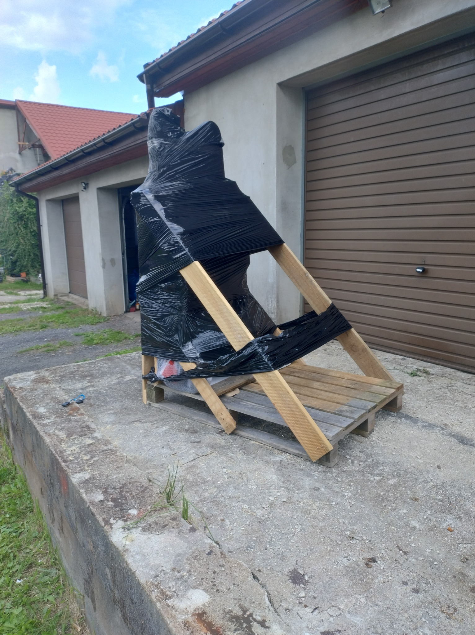 Element zabezpieczony czarną folią i drewnianą konstrukcją, prawdopodobnie do transportu lub przechowywania, na zewnątrz budynku z garażami.