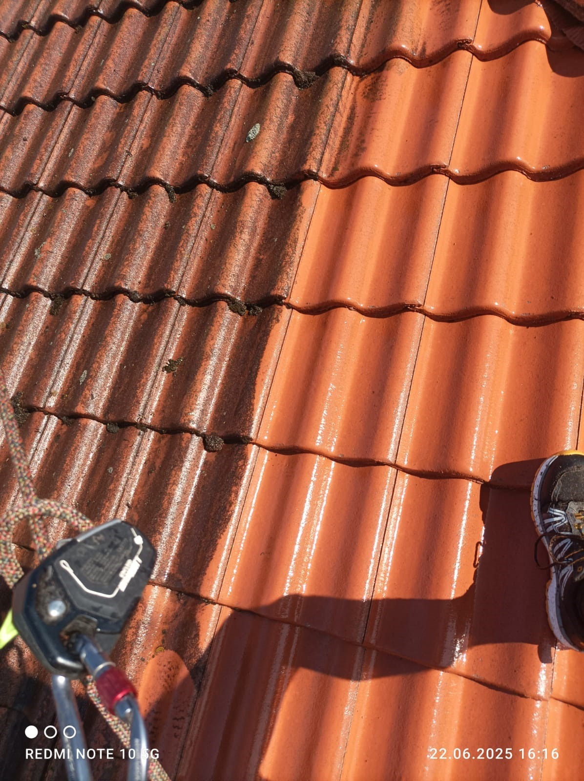 Częściowo wyczyszczona, czerwona dachówka z wyraźną różnicą w kolorze, z widocznymi śladami wody, sprzęt alpinistyczny i fragment buta w kadrze.
