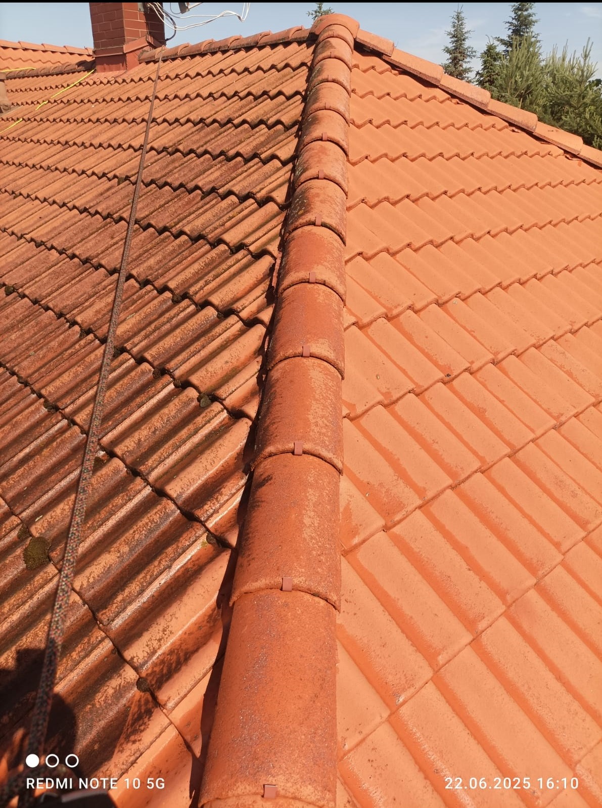 Częściowo umyty dach z czerwonej dachówki ceramicznej, widoczne zabrudzenia po lewej stronie i czysta powierzchnia po prawej. Perspektywa z góry, widoczny fragment liny.