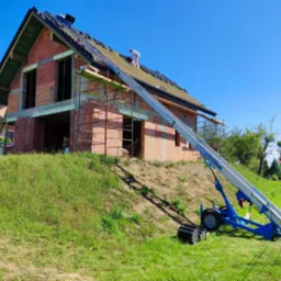 Budowa domu z czerwonej cegły, widoczny fragment dachu z nową dachówką, rusztowania i podnośnik dekarski na tle błękitnego nieba.