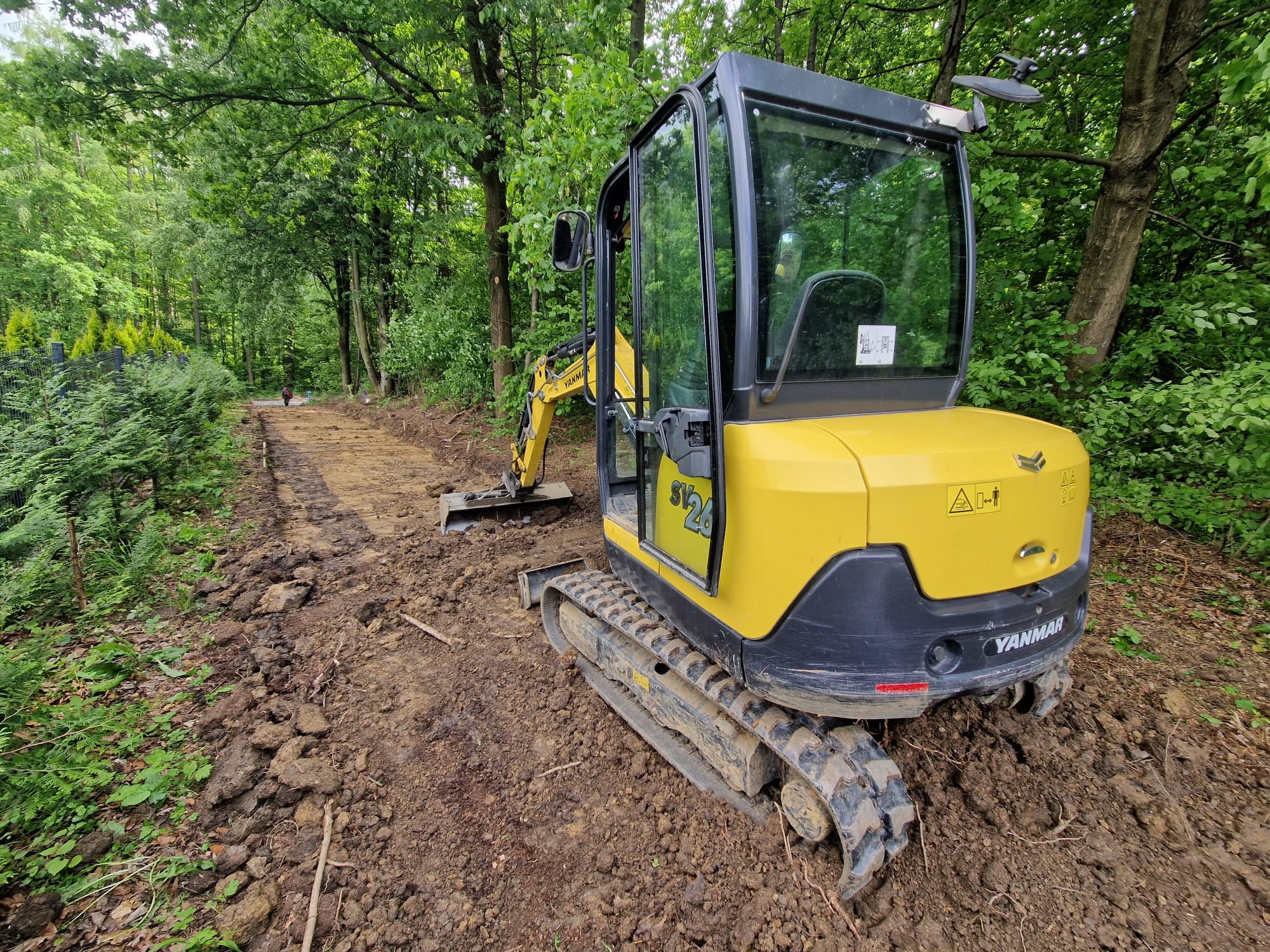 Żółta mini koparka Yanmar SV26 na terenie budowy w lesie, przygotowująca teren pod ścieżkę. Widoczne świeżo przekopane błoto i fragment wyrównanego terenu.