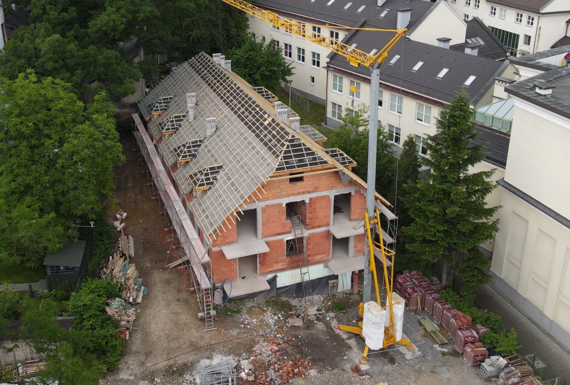 Budowa domu z czerwonej cegły, widok z góry. Częściowo wykonane krycie dachu z deskowaniem i papą. Na budowie stoi żółty dźwig, widoczne rusztowania.