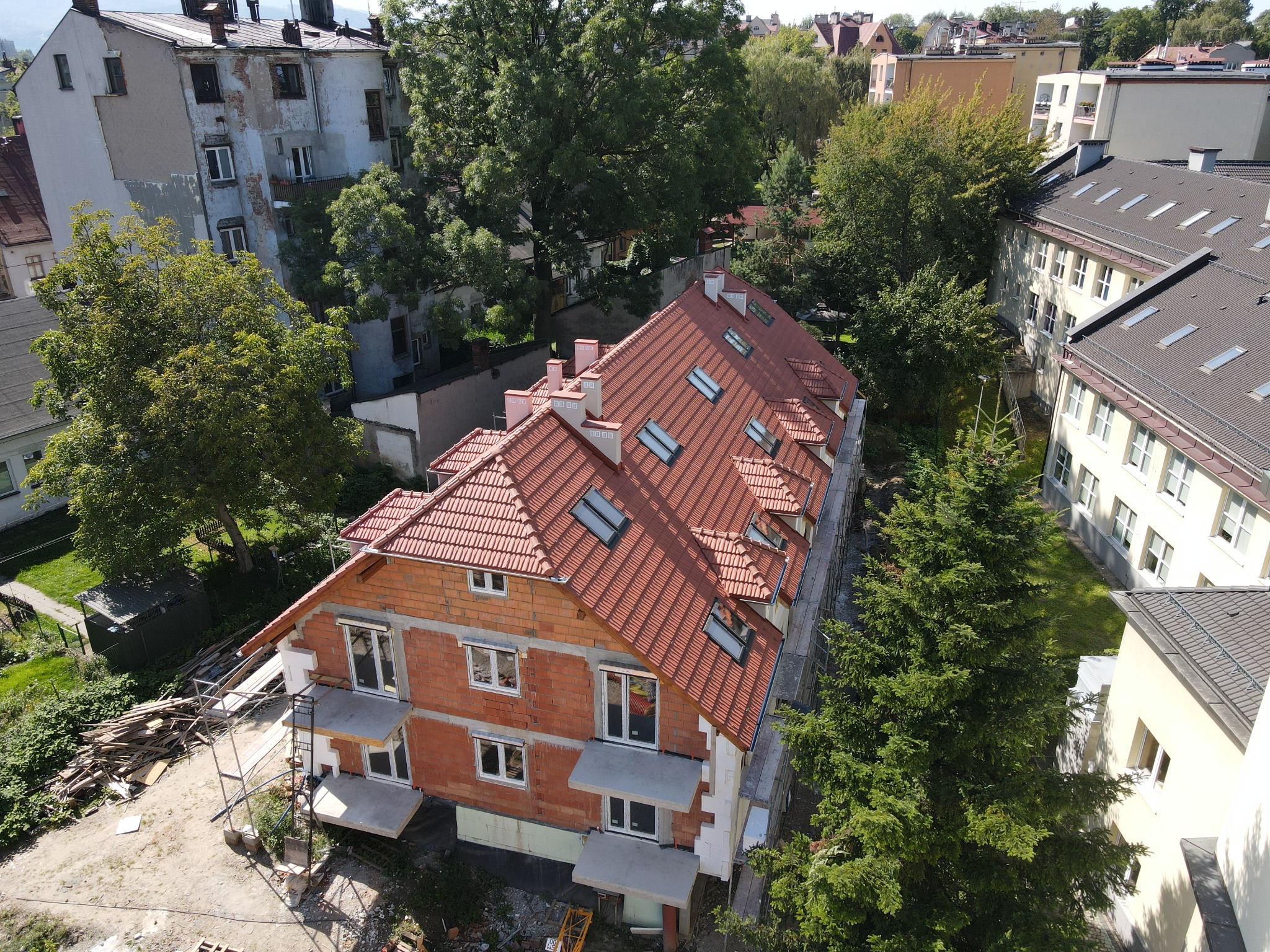 Ceglany budynek w trakcie budowy z czerwonym dachem i oknami dachowymi, widziany z góry, otoczony zielenią i innymi budynkami. Widoczne rusztowania i materiały budowlane.