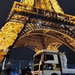 PUBLICZNY TRANSPORT DROGOWY CIĘŻAROWY BOGDAN STYPCZYŃSKI - Ciężarówka na tle wieży Eiffla w Paryżu nocą. Transport z logo firmy. Częściowo widoczni ludzie. Ujęcie z dołu, oświetlona wieża.