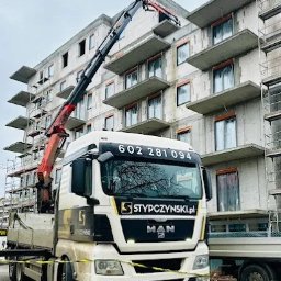 PUBLICZNY TRANSPORT DROGOWY CIĘŻAROWY BOGDAN STYPCZYŃSKI - Ciężarówka MAN z dźwigiem firmy Stypczynski pod budynkiem w trakcie budowy. Dźwig podnosi element na wyższe piętro. Widoczne numery kontaktowe na kabinie.