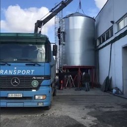 PUBLICZNY TRANSPORT DROGOWY CIĘŻAROWY BOGDAN STYPCZYŃSKI - Niebieska ciężarówka z dźwigiem podnosi srebrny silos zbożowy na tle błękitnego nieba z chmurami, montaż silosów, pracownicy na drabinie i dachu budynku.