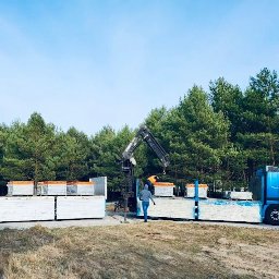 PUBLICZNY TRANSPORT DROGOWY CIĘŻAROWY BOGDAN STYPCZYŃSKI - Niebieska ciężarówka z dźwigiem załadowana płytami budowlanymi na tle lasu. Prace załadunkowe w toku, widoczny pracownik w odblaskowej kamizelce.