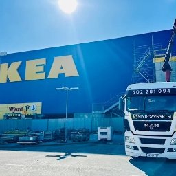 PUBLICZNY TRANSPORT DROGOWY CIĘŻAROWY BOGDAN STYPCZYŃSKI - Ciężarówka MAN z wysięgnikiem na tle budynku IKEA w słoneczny dzień. Na dachu budynku pracownicy w pomarańczowych kamizelkach. Transport materiałów budowlanych.