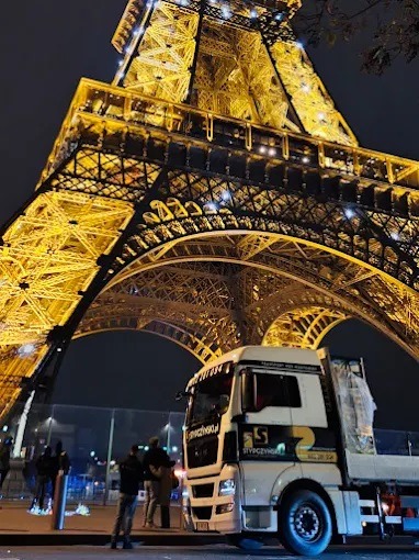Ciężarówka na tle wieży Eiffla w Paryżu nocą. Transport z logo firmy. Częściowo widoczni ludzie. Ujęcie z dołu, oświetlona wieża.
