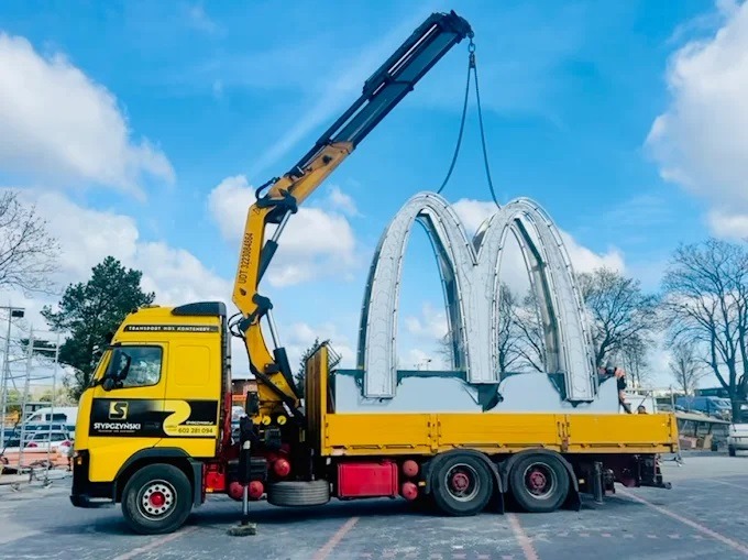 Żółta ciężarówka z dźwigiem transportuje metalowe logo McDonald's na niebieskim niebie. Widoczny napis 'Transport HDS kontenery' i logo firmy przewozowej.