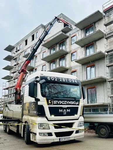 Ciężarówka MAN z dźwigiem firmy Stypczynski pod budynkiem w trakcie budowy. Dźwig podnosi element na wyższe piętro. Widoczne numery kontaktowe na kabinie.
