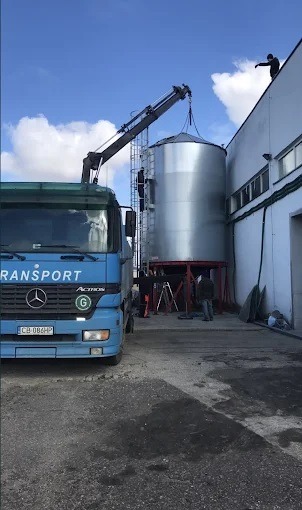 Niebieska ciężarówka z dźwigiem podnosi srebrny silos zbożowy na tle błękitnego nieba z chmurami, montaż silosów, pracownicy na drabinie i dachu budynku.