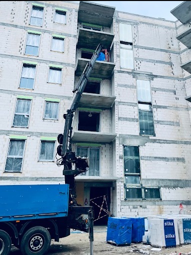 Żuraw samochodowy podnosi materiały budowlane na balkon powstającego bloku z białej cegły. W tle widoczne okna z roletami i balkony.