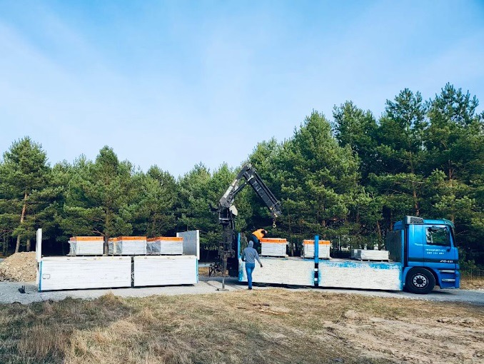 Niebieska ciężarówka z dźwigiem załadowana płytami budowlanymi na tle lasu. Prace załadunkowe w toku, widoczny pracownik w odblaskowej kamizelce.