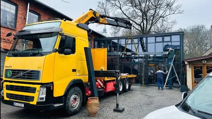 Żółta ciężarówka z dźwigiem podnosi element konstrukcyjny przeszklonej oranżerii. Dwóch monterów na drabinie montuje element. Widok na budynek z cegły i drzewa.