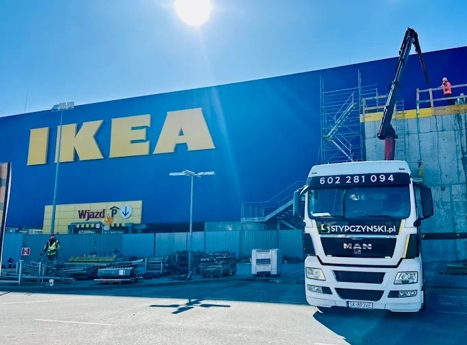 Ciężarówka MAN z wysięgnikiem na tle budynku IKEA w słoneczny dzień. Na dachu budynku pracownicy w pomarańczowych kamizelkach. Transport materiałów budowlanych.