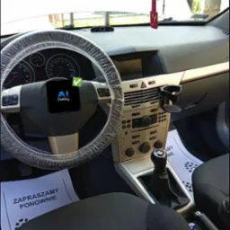 Wnętrze samochodu z pokrowcem na kierownicy i kartkami 'Zapraszamy ponownie' na dywanikach, sugerujące świeże czyszczenie tapicerki.