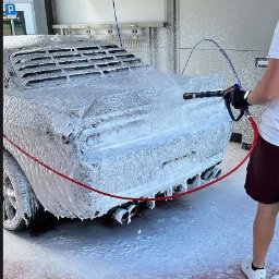 AI Detailing - Luksusowy samochód pokryty pianą podczas mycia pod ciśnieniem w myjni. Widoczne dysze i czerwony wąż. Czerwona podłoga myjni. Ujęcie z boku, widok na tył auta.