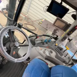 Wnętrze kabiny traktora z niebieskimi siedzeniami i kierownicą w folii ochronnej. Widoczny kokpit z wyświetlaczami i dźwigniami. Ujęcie od dołu, lekko z ukosa.