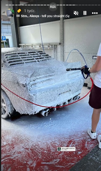 Luksusowy samochód pokryty pianą podczas mycia pod ciśnieniem w myjni. Widoczne dysze i czerwony wąż. Czerwona podłoga myjni. Ujęcie z boku, widok na tył auta.