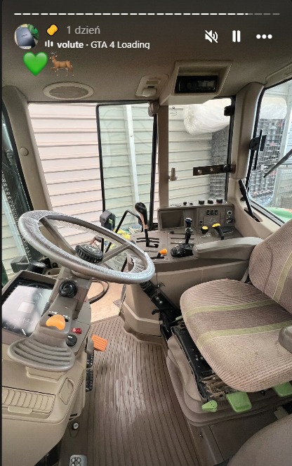 Wnętrze kabiny ciągnika John Deere z widoczną kierownicą, siedzeniem operatora i panelem sterowania. Jasne wnętrze z elementami zieleni i beżu.