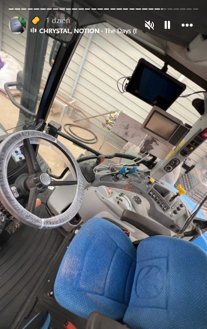 Wnętrze kabiny traktora z niebieskimi siedzeniami i kierownicą w folii ochronnej. Widoczny kokpit z wyświetlaczami i dźwigniami. Ujęcie od dołu, lekko z ukosa.