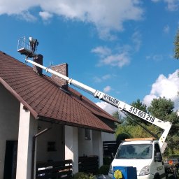 Drew-Wart - Pracownik w koszu podnośnika na dachu domu jednorodzinnego z brązową dachówką. Podnośnik z napisem 'Wynajem' i numerem telefonu. Błękitne niebo z chmurami w tle.