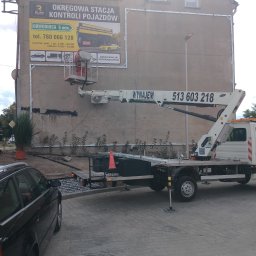 Drew-Wart - Pracownik na podnośniku koszowym montuje ramę pod reklamę na elewacji budynku. Widoczny samochód i fragment chodnika z włazem.