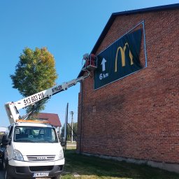 Drew-Wart - Montaż reklamy McDonald's na ceglanej ścianie budynku za pomocą podnośnika koszowego na tle błękitnego nieba i drzewa.