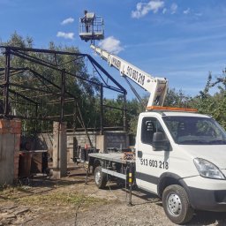 Drew-Wart - Podnośnik koszowy z pracownikiem na tle metalowej konstrukcji budynku. Pojazd z wysięgnikiem na nierównym terenie, widoczne drzewa i błękitne niebo.