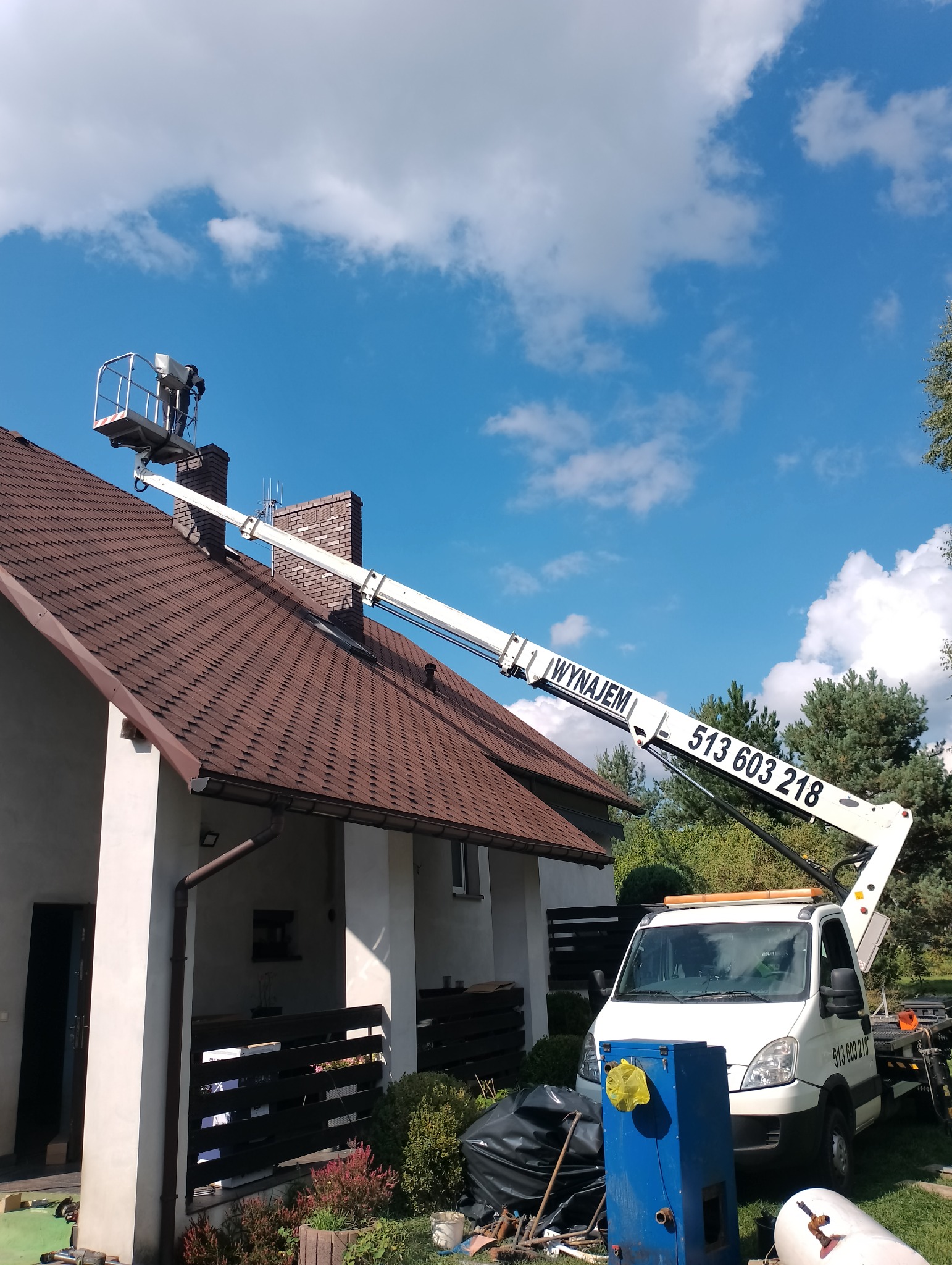 Pracownik w koszu podnośnika na dachu domu jednorodzinnego z brązową dachówką. Podnośnik z napisem 'Wynajem' i numerem telefonu. Błękitne niebo z chmurami w tle.