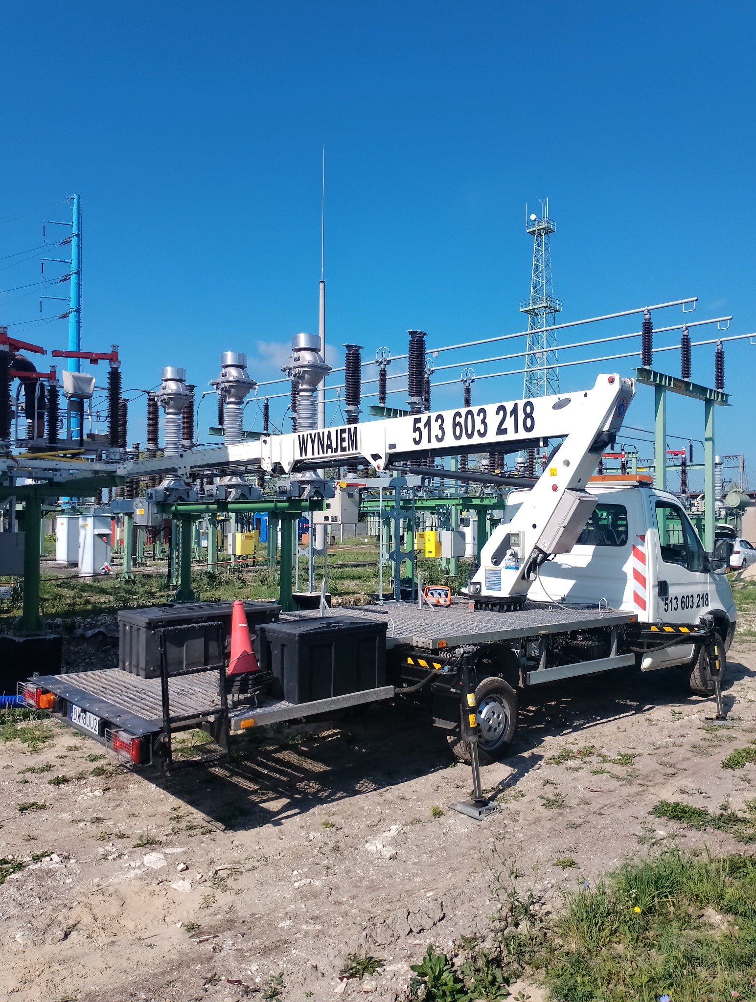 Podnośnik koszowy na przyczepie z numerem telefonu, ustawiony na terenie stacji transformatorowej, błękitne niebo w tle.