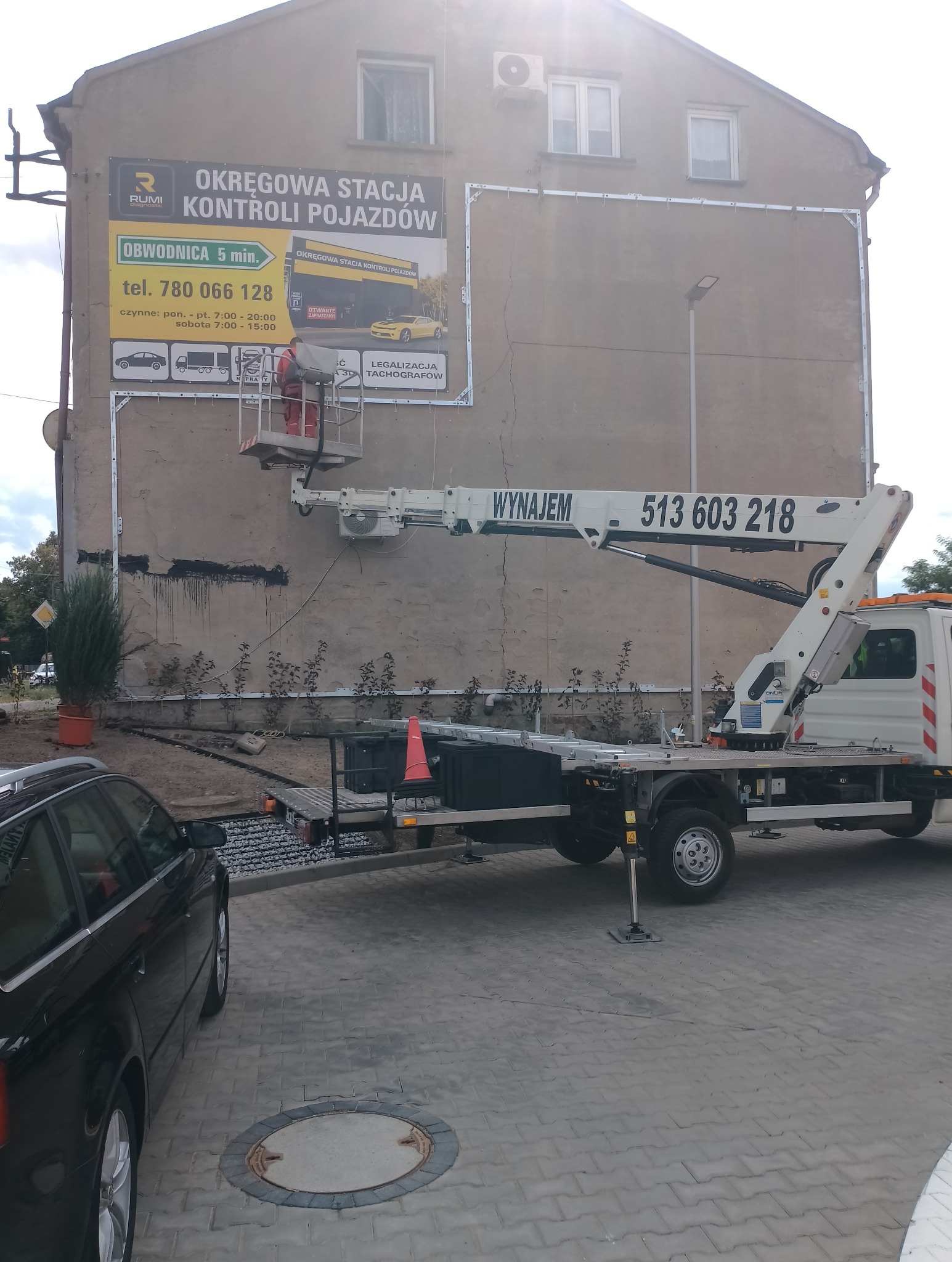 Pracownik na podnośniku koszowym montuje ramę pod reklamę na elewacji budynku. Widoczny samochód i fragment chodnika z włazem.