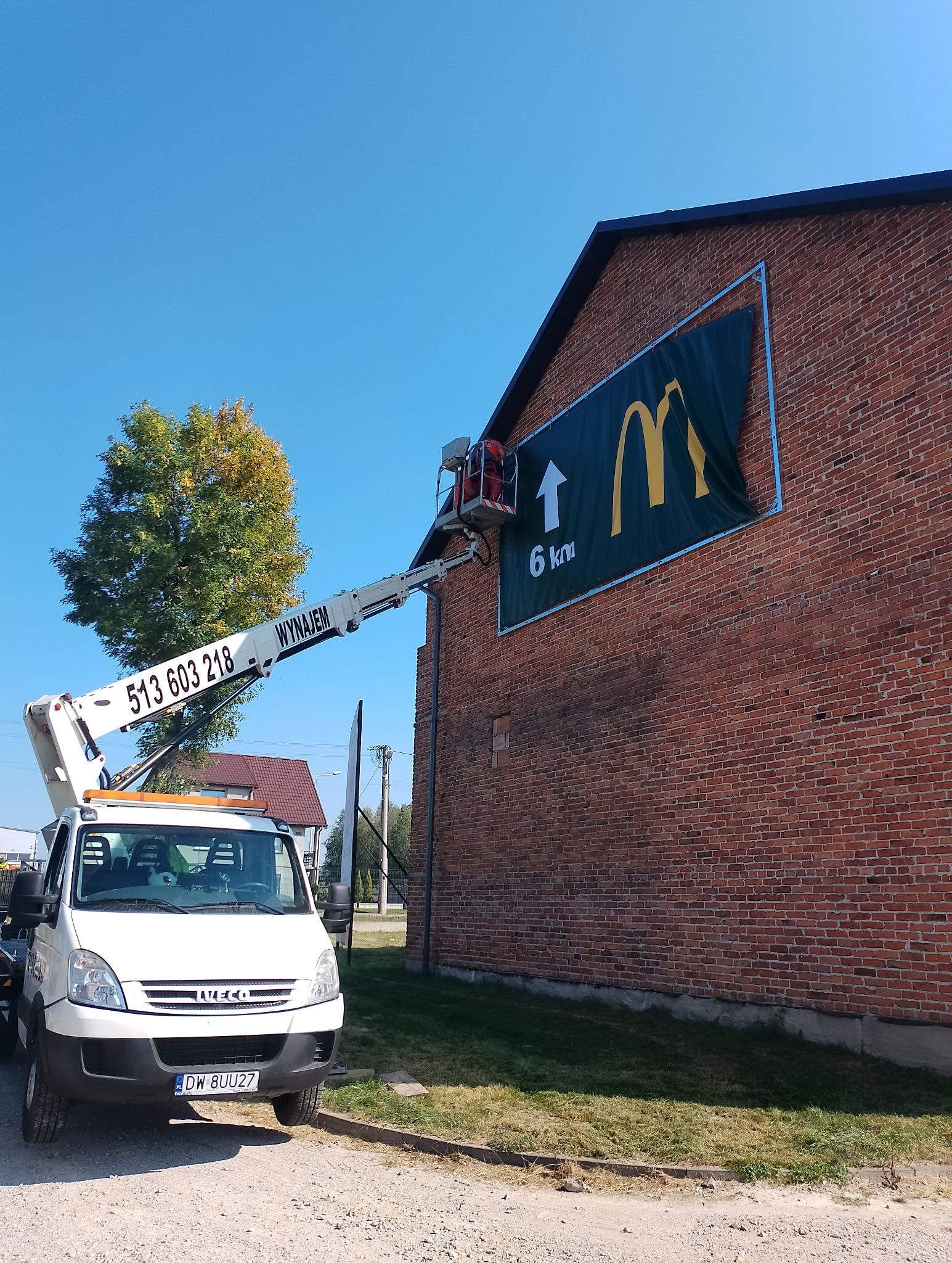 Montaż reklamy McDonald's na ceglanej ścianie budynku za pomocą podnośnika koszowego na tle błękitnego nieba i drzewa.