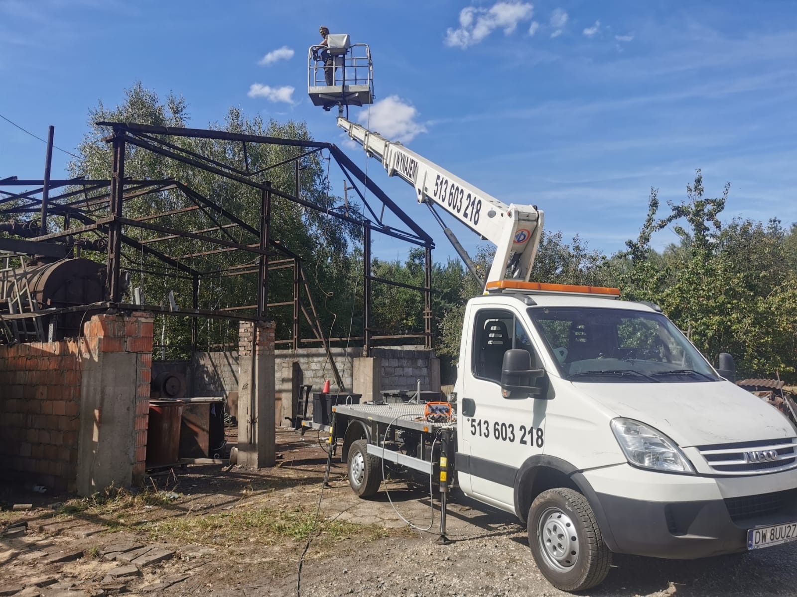 Podnośnik koszowy z pracownikiem na tle metalowej konstrukcji budynku. Pojazd z wysięgnikiem na nierównym terenie, widoczne drzewa i błękitne niebo.