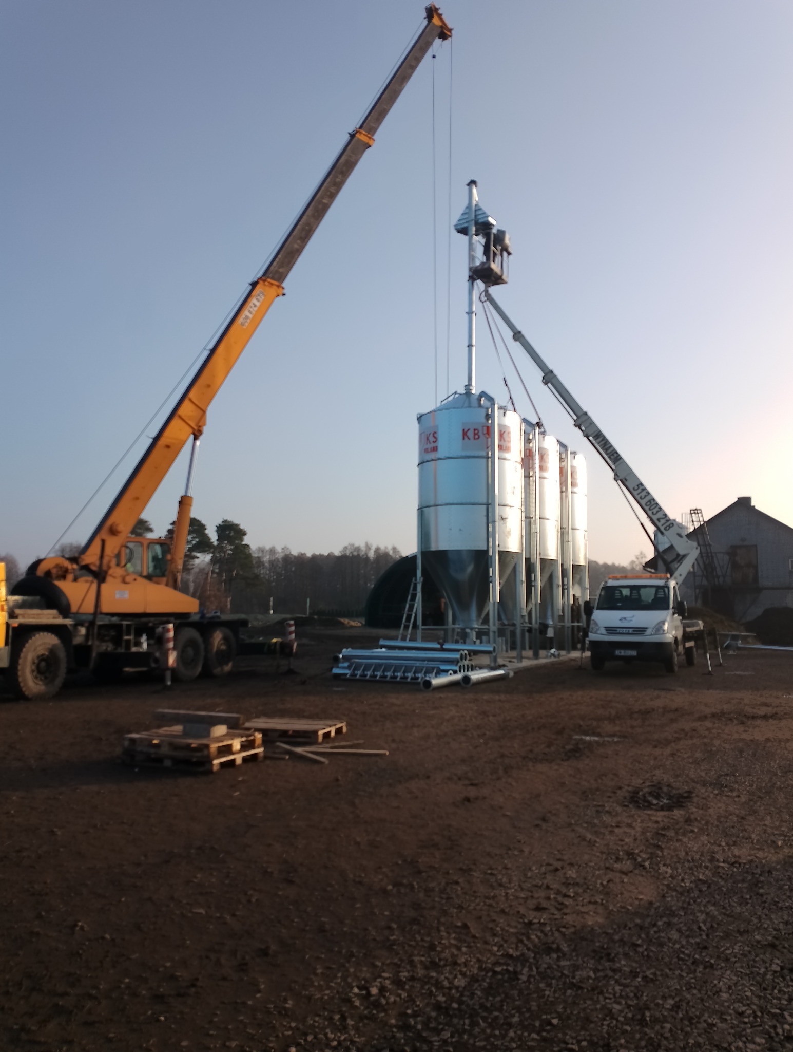 Montaż silosów zbożowych z użyciem dźwigu i podnośnika koszowego na placu budowy. Widoczne elementy konstrukcyjne i sprzęt ciężki.