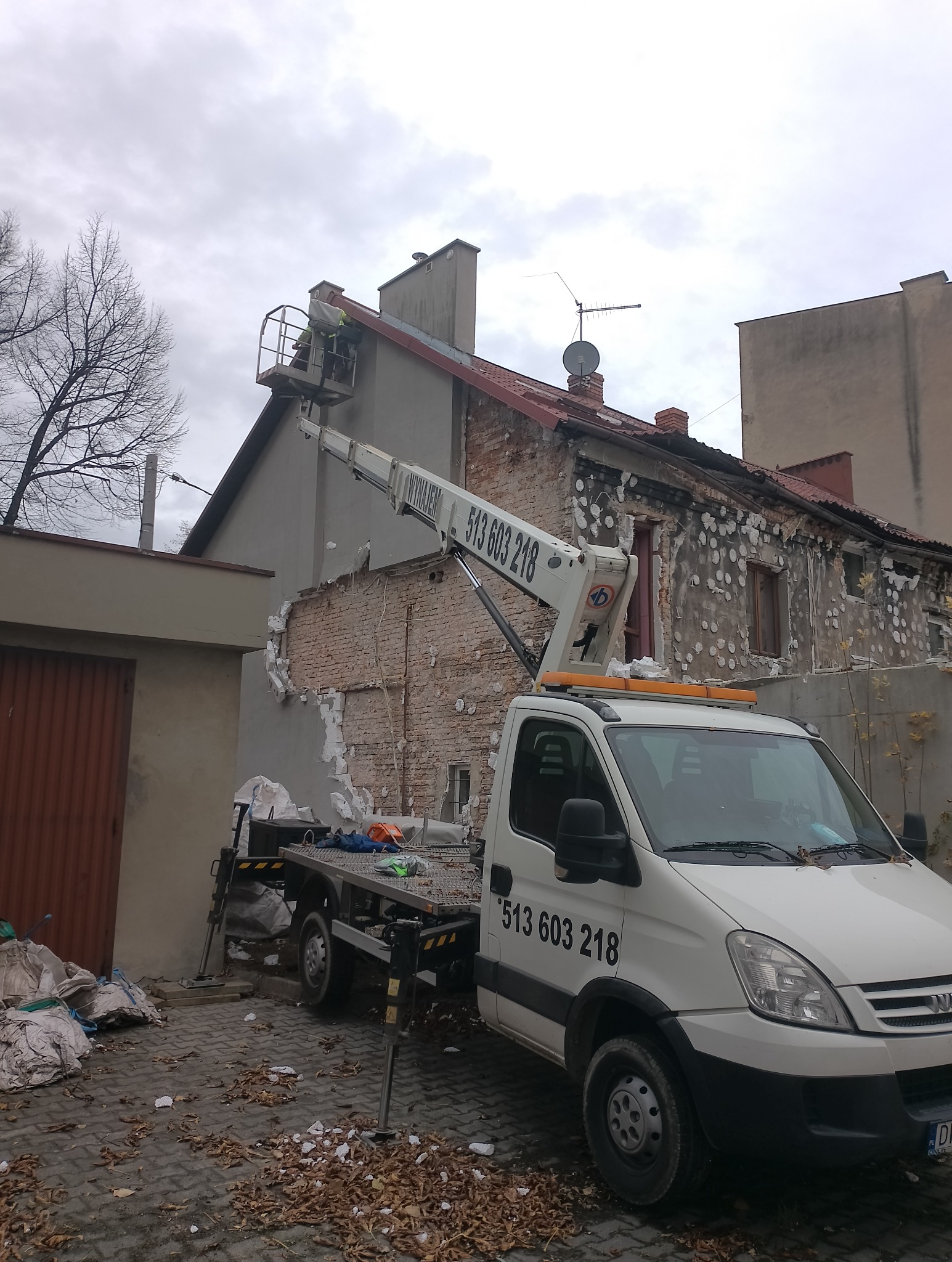 Pracownik na platformie podnośnika koszowego, demontujący ocieplenie elewacji budynku. Widoczny fragment odsłoniętej cegły i gruz na ziemi. Numer kontaktowy na pojeździe.