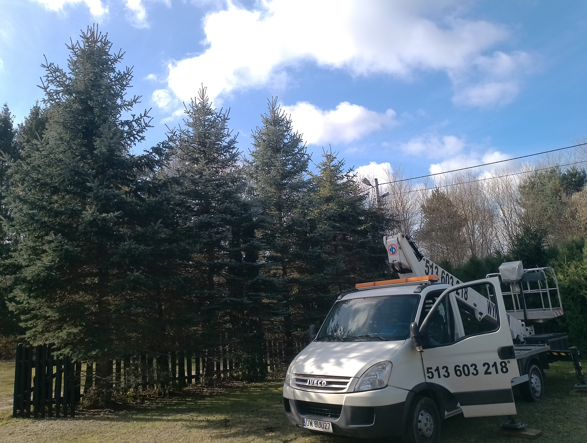 Biały samochód z podnośnikiem koszowym na tle wysokich, zielonych świerków i błękitnego nieba z chmurami. Widoczny numer telefonu na pojeździe.