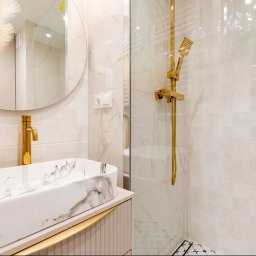 KLIMAR MICHAŁ CHMIEL - Elegancka łazienka po remoncie: marmurowa umywalka, złota armatura, kabina prysznicowa z mozaiką podłogową i okrągłe lustro. Nowoczesny design z akcentem roślinnym.