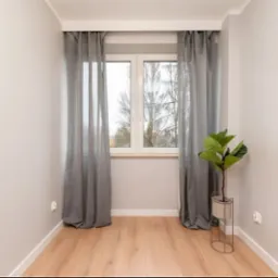 Jasne wnętrze po remoncie: świeżo pomalowane ściany, drewniana podłoga, okno z szarymi zasłonami i roślina w minimalistycznej donicy.