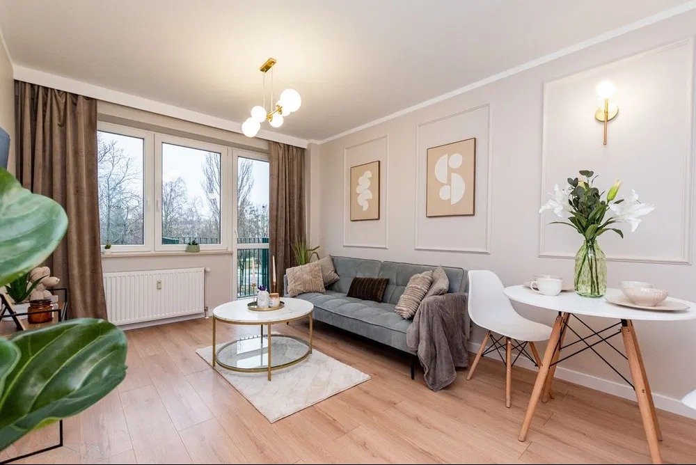 Jasne wnętrze salonu po remoncie: drewniana podłoga, szara sofa, białe ściany z dekoracyjnymi ramami, okno z brązowymi zasłonami, okrągły stolik i designerskie lampy.