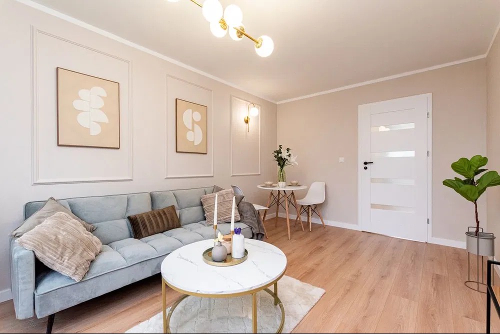 Jasny salon po remoncie: błękitna sofa, okrągły stolik z marmurowym blatem, drewniana podłoga, obrazy na ścianach w kremowych ramach i nowoczesne oświetlenie.