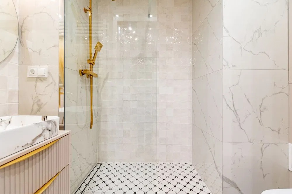 Wykończona łazienka z kabiną prysznicową typu walk-in, marmurowymi płytkami na ścianach i wzorzystą podłogą. Złota armatura i umywalka z marmurowym wzorem.