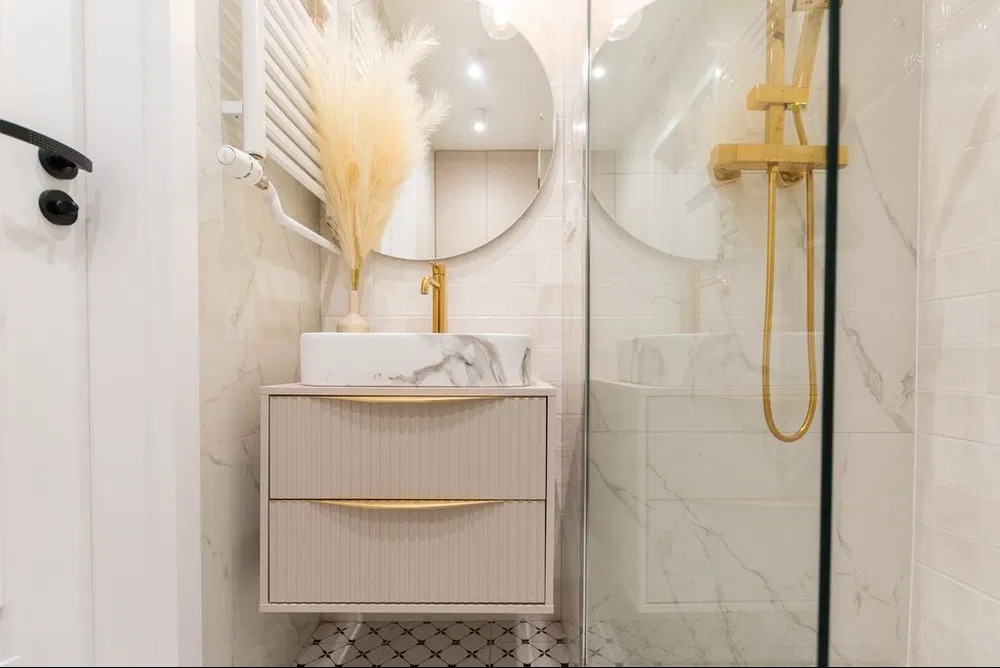 Nowoczesna łazienka po remoncie: marmurowa umywalka na szafce z frezowanymi frontami, złota armatura, okrągłe lustro i szklana kabina prysznicowa.