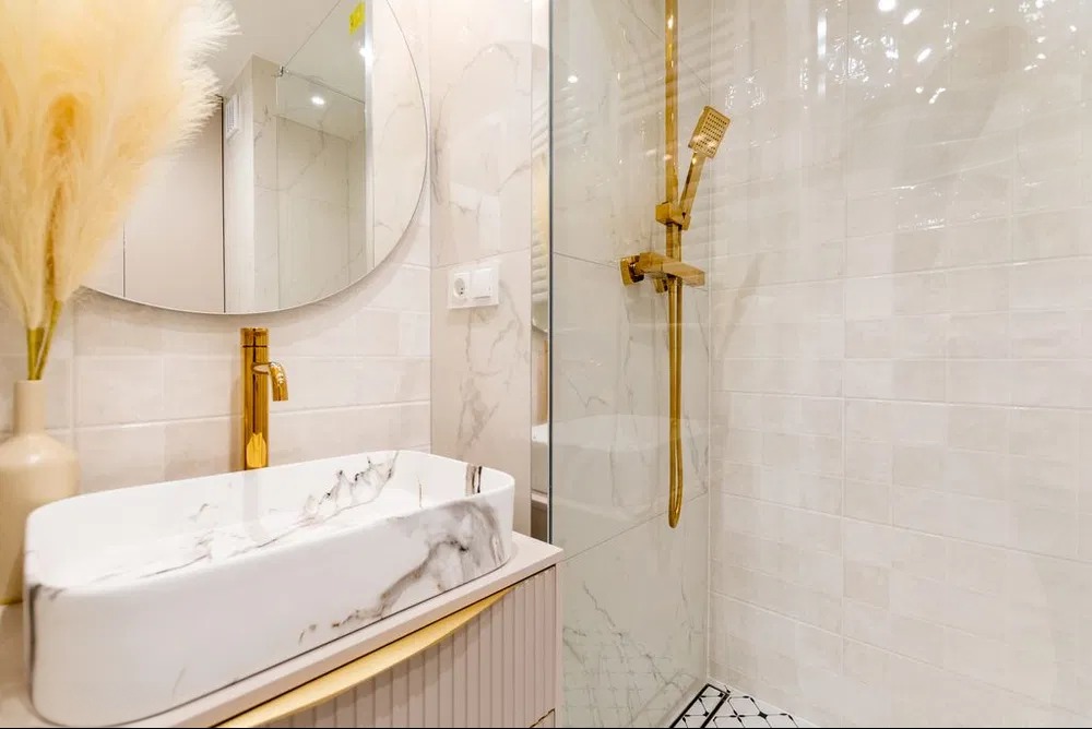 Elegancka łazienka po remoncie: marmurowa umywalka, złota armatura, kabina prysznicowa z mozaiką podłogową i okrągłe lustro. Nowoczesny design z akcentem roślinnym.