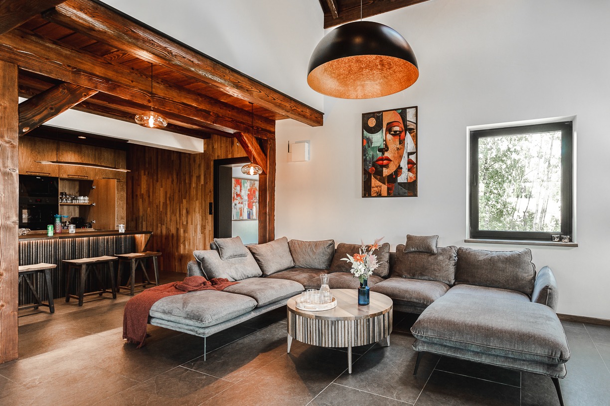Salon z drewnianymi belkami stropowymi, szarą sofą narożną i okrągłym stolikiem kawowym. Na ścianie wisi abstrakcyjny obraz, a nad sofą lampa w kształcie kuli.