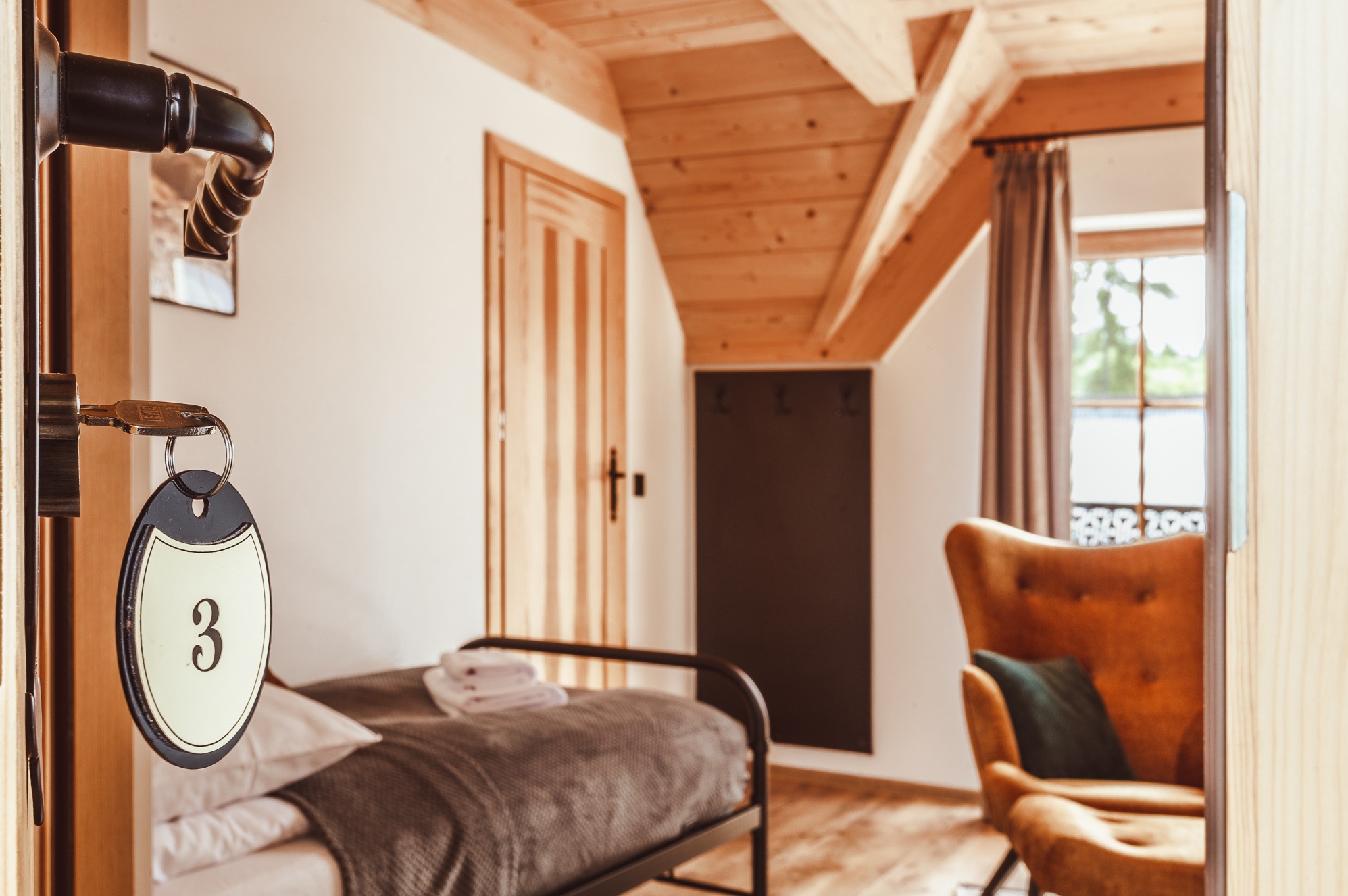 Klucz z numerem 3 w drzwiach pokoju hotelowego z drewnianym sufitem, łóżkiem i fotelem w tle. Ciepłe oświetlenie i przytulna atmosfera.